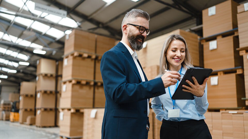 male and female factory distribution center managers reviewing shipment data on an electronic device inside the warehouse