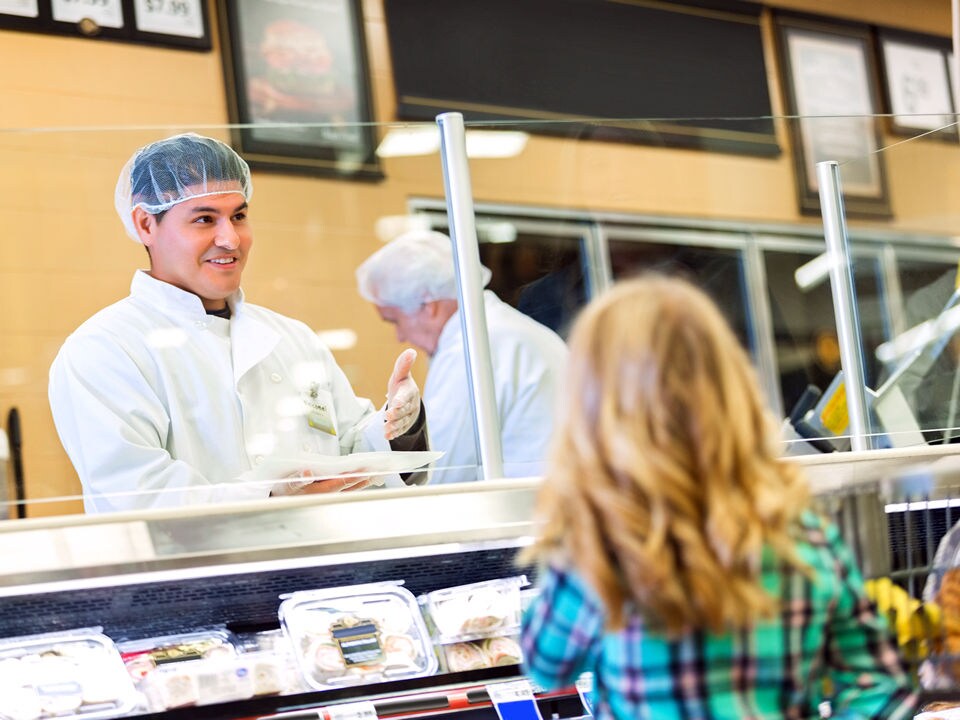 a male grocery deli counter employee in sanitary hair net and white butcher coat stands behind the counter cold case and helps a customer