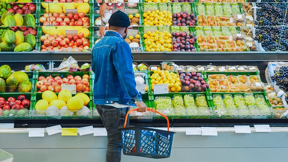 middle age man wearing blue jacket and knit cap carrying a shopping basket in retail grocery store in fornt of array of fresh fruits and vegetables