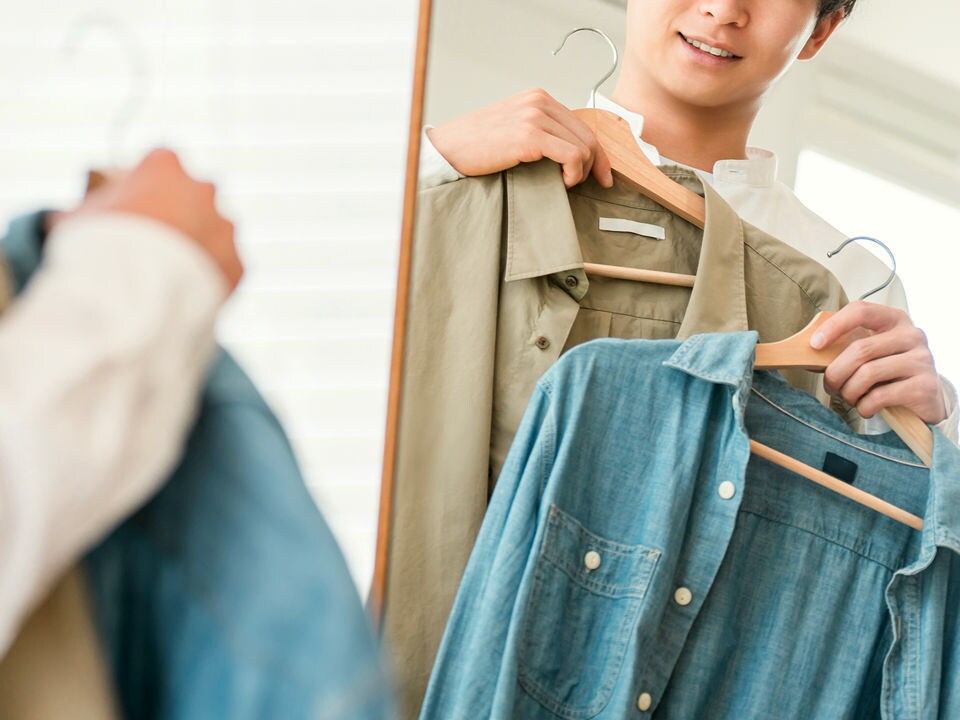 young adult shopper standing in front of mirror holding up two shirt against their body in retail apparel store fitting room