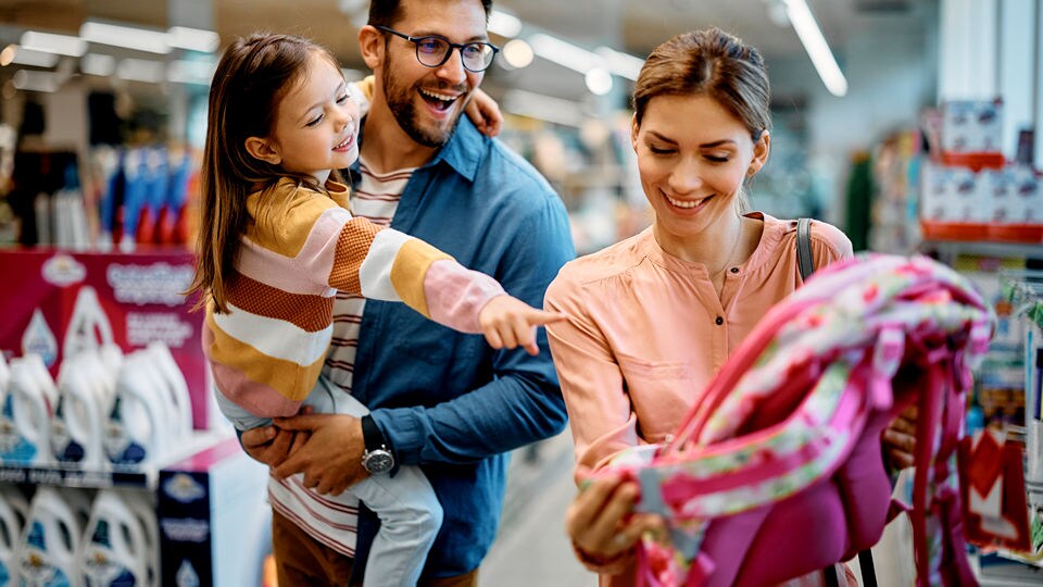 father holds young daughter pointing at a backpack held by her mother which she likes in a retail department store