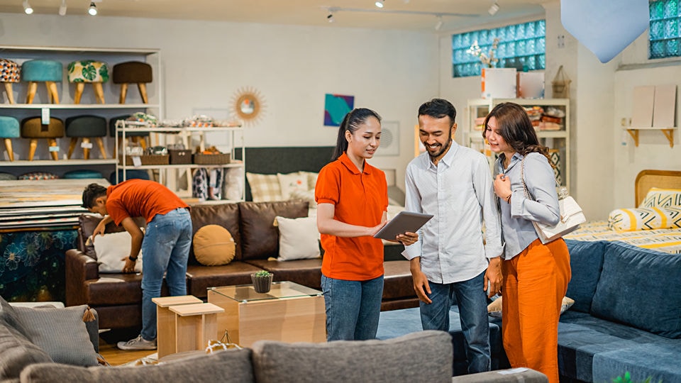 male and female shoppers in a retail furniture store are speaking with a sales associate holding an electronic tablet while another associate in the background tidies up the showroom