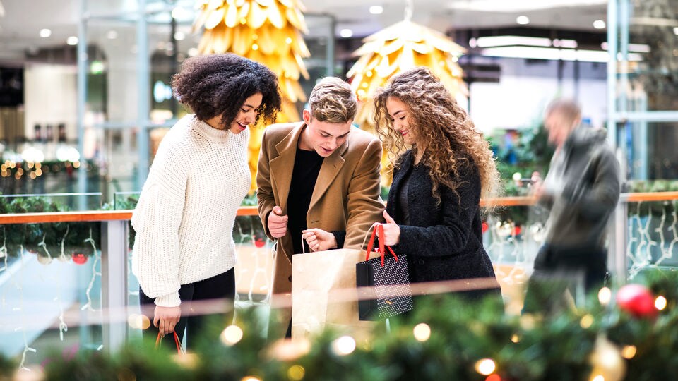 friends group of 2 women and 1 man looking over their hliday purchases in shopping bags at retail mall decorated for holiday season