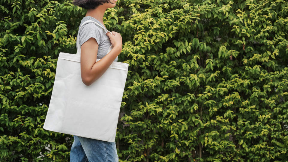 woman wearing jeans and t-shirt carrying cloth shopping bag in front of greenery