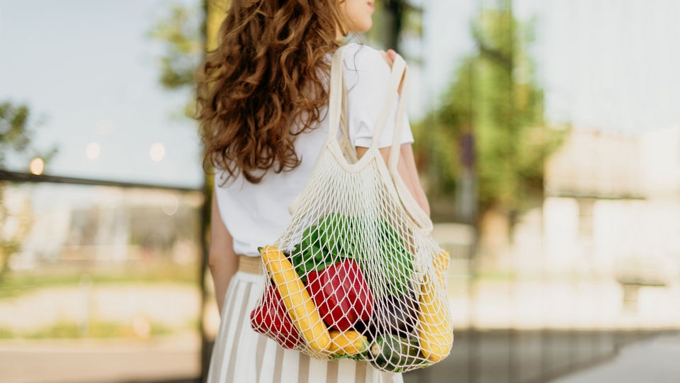 woman in white dress walking on tree-lined street carrying cloth mesh recyclable shopping bag with fruits and vegetables