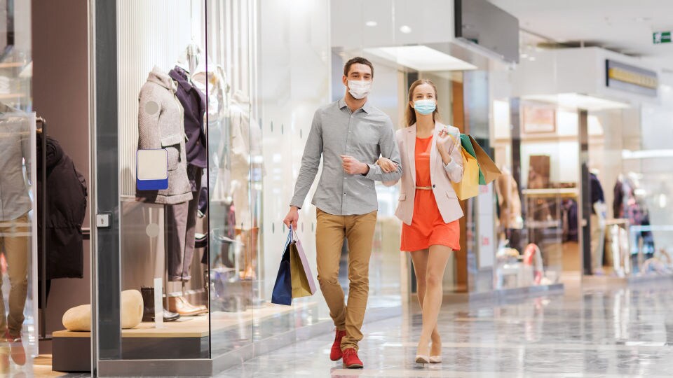 masked male and female shoppers walking through mall carrying purchases