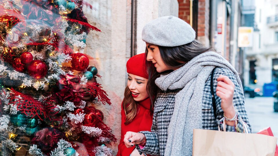 two women shoppers dressed for cold weather looking into a retail store holiday decorated window on a shopping street