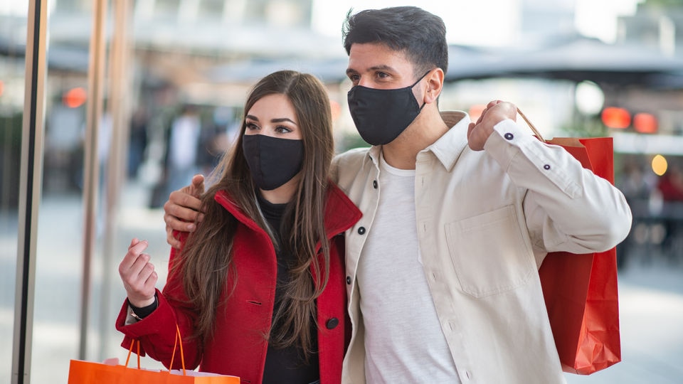male and female retail shoppers wearing masks look in store window while carrying shopping bags with purchases