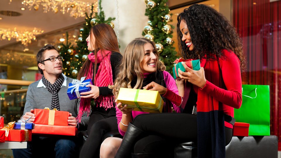 group of friends including one male and three females resting in retail shopping mall decorated for holidays, holding wrapped gifts and shopping bags 