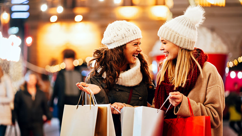 two women shoppers dressed for cold weather are talking and carrying multiple purchases in shopping bags through retail shopping center