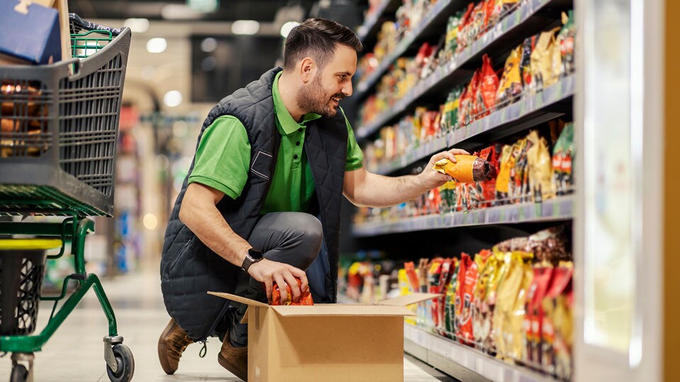 a male grocery store employee is kneeling in the aisle of the store putting stock on the shelf