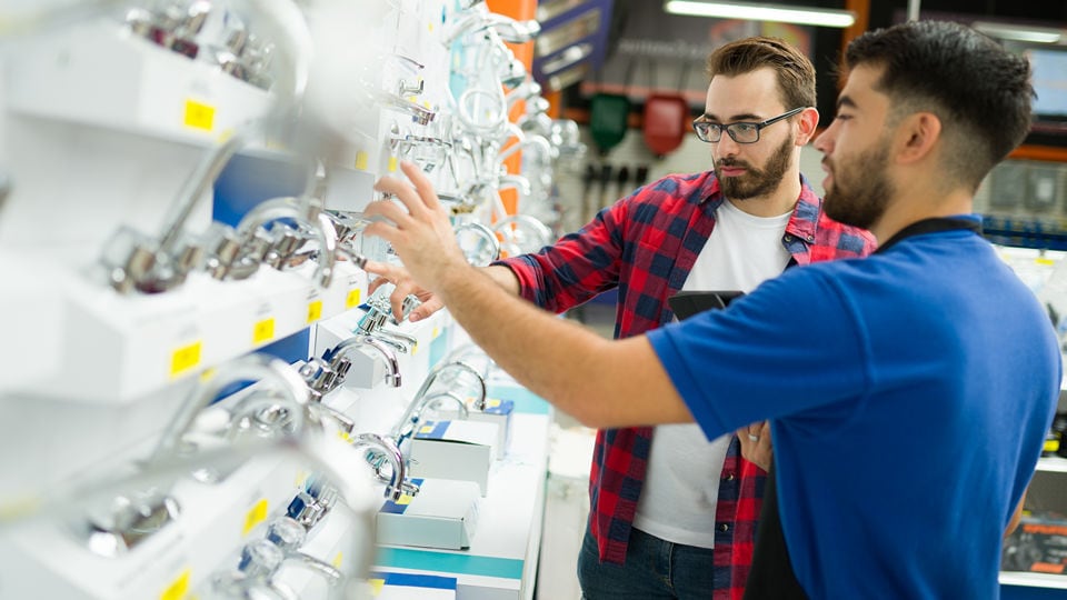 male shopper with beard and glasses is helped by a young male sales associate at the plumbing fixture display in a retail hardware store