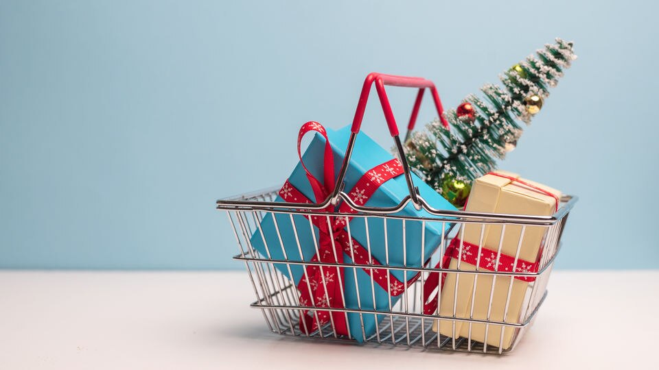 handheld wire retail shopping basket containing holiday wrapped gifts and a miniature decorated christmas tree