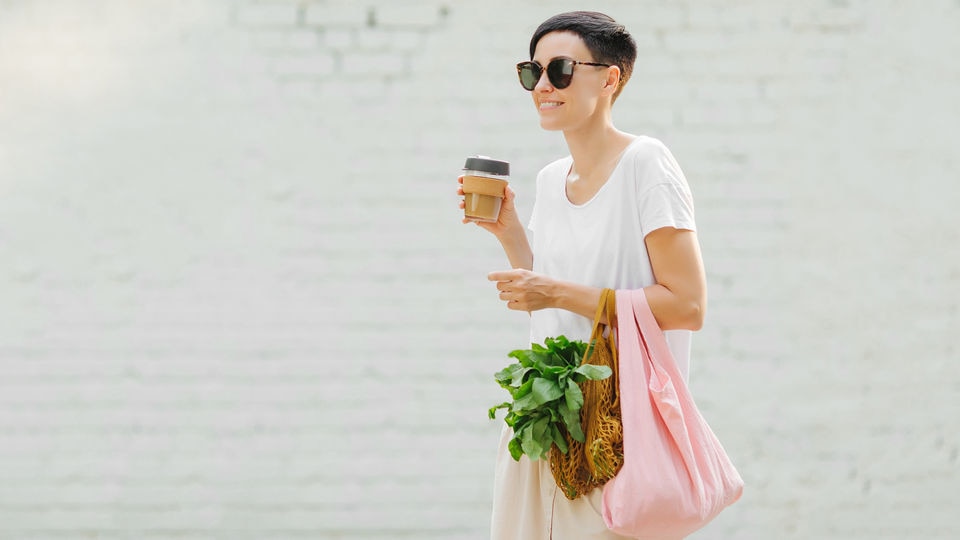 woman in sunglasses carrying environmentally-friendly coffee cup and shopping bag from retail grocery
