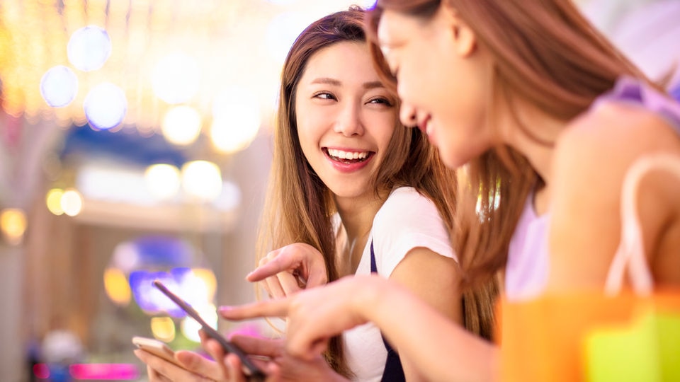 two young Asian female shoppers looking at their smartphones in a retail store.