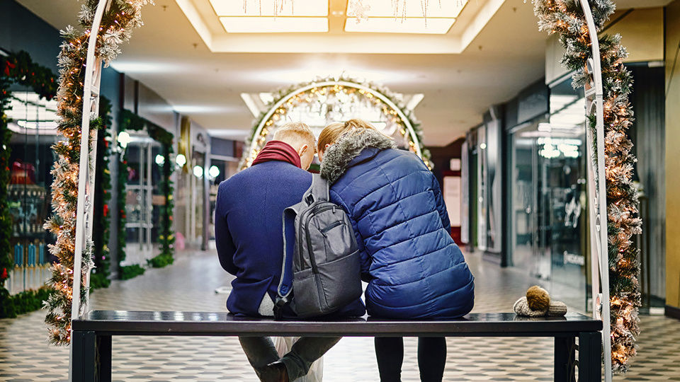 male and female couple in winter outerwear enjoying cozy moment sitting on a bench in a holiday decorated shopping mall