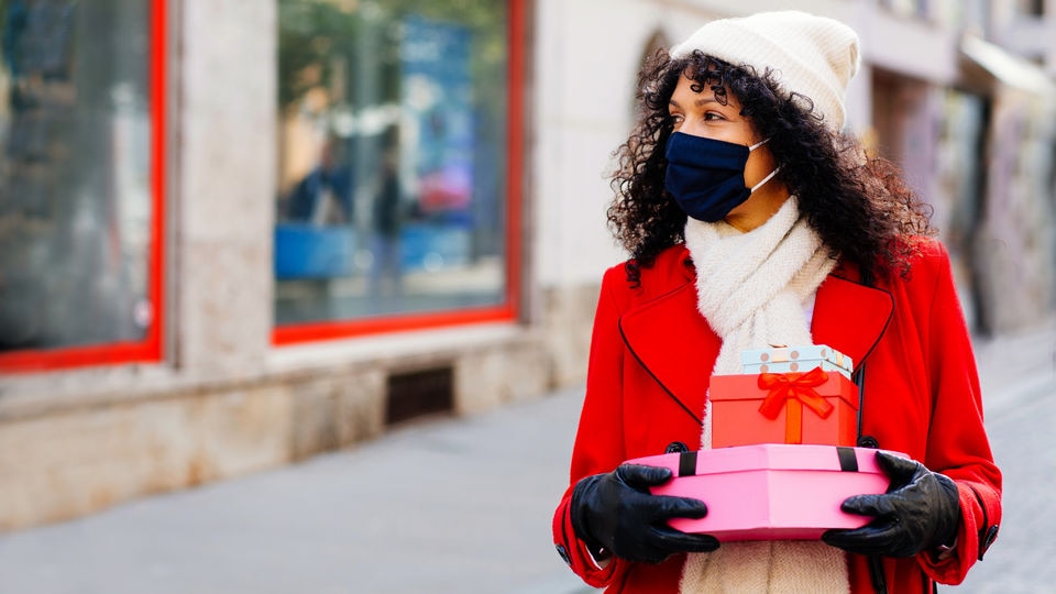 female shopper wearing mask and red winter coat carrying wrapped gifts on shopping street in front of retail store