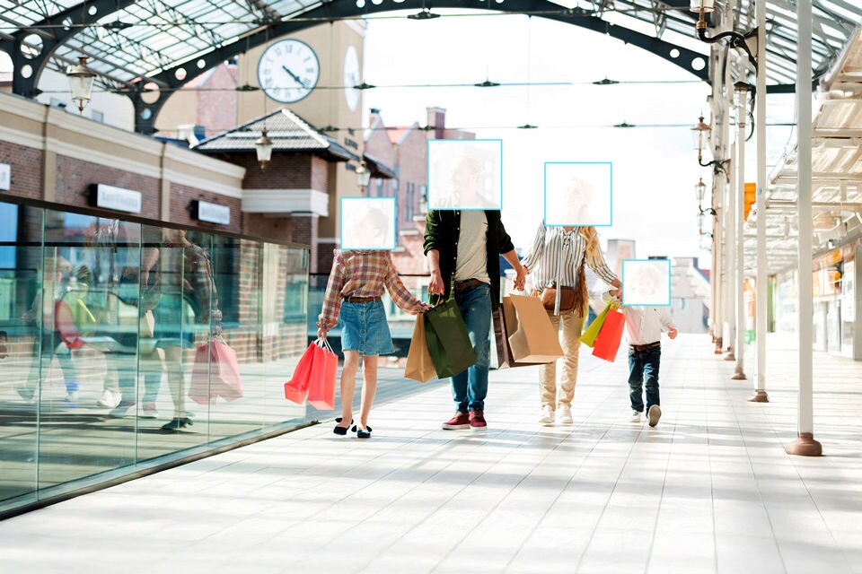 father mother and two children holding hands while carrying shopping bags through glassroofed outdoor shopping mall with their faces covered by simulated screens