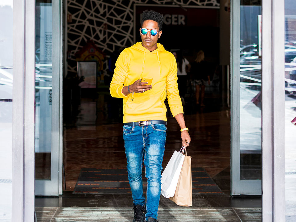 stylish young man in sunglasses yellow sweater and jeans leaving retail store carrying shopping bag holding cellphone