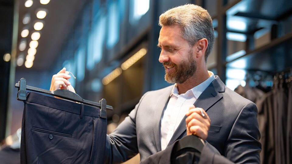 senior male shopper with silver hair and beard choosing trousers and a sport jacket at a mens apparel retail store