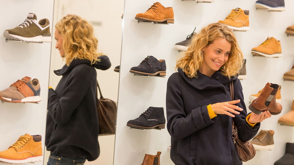 young female shopper looking at shoes on a display wall in a retail shoe store with her back reflected in a mirror