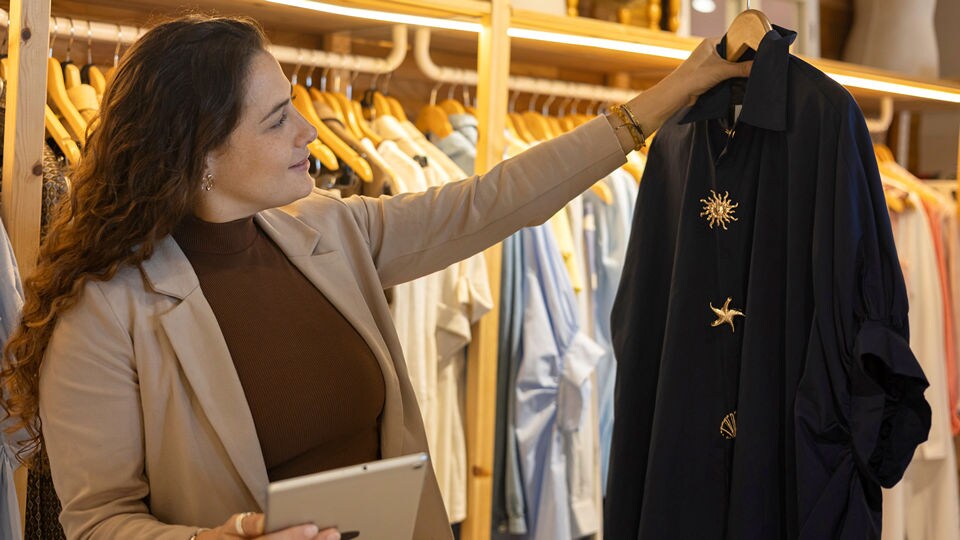 female retail associate in a womens apparel store holding an electronic tablet in one hand holding up a stylish jacket in the other