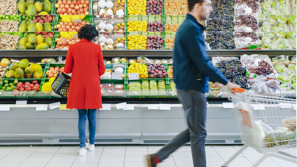 woman browsing in retail grocery produce aisle while man with shopping cart passes in the foreground