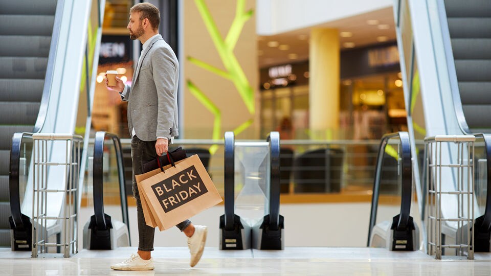 stylish man walks in busy retail shopping mall carrying a cup of coffee and a shopping bag with black friday logo