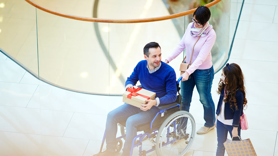 disabled man in wheelchair holds a gift box purchase while his wife and daughter accompany him through retail shopping mall