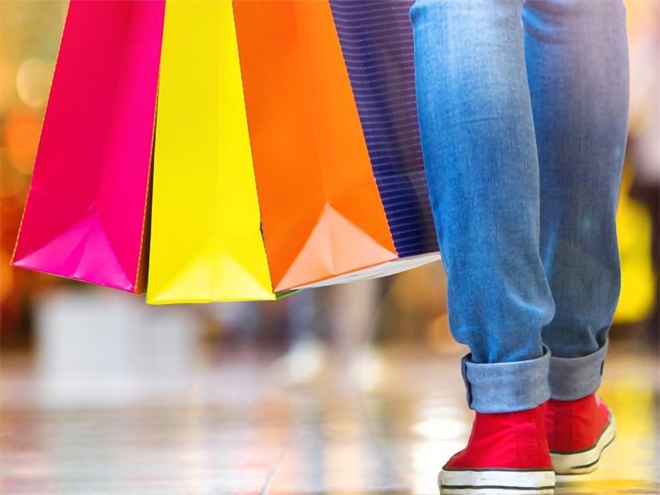 shoppers legs and feet in jeans and red sneakers carrying several retail shopping bags in bright colors through shopping mall