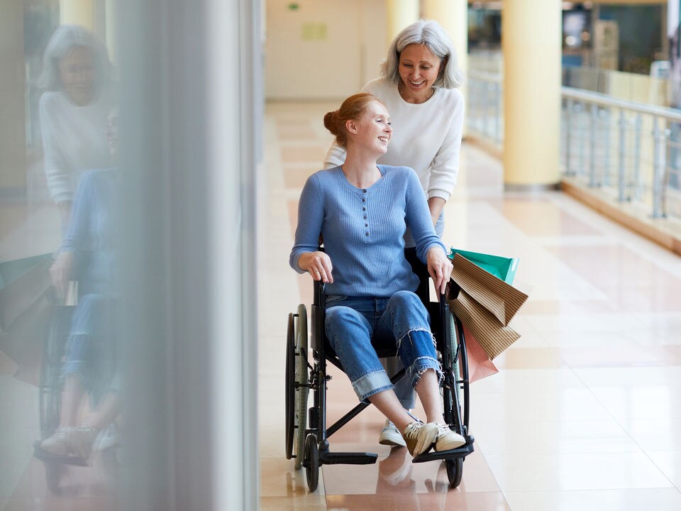 older female pushing younger female in a wheelchair while carrying shopping bags through a retail shopping mall