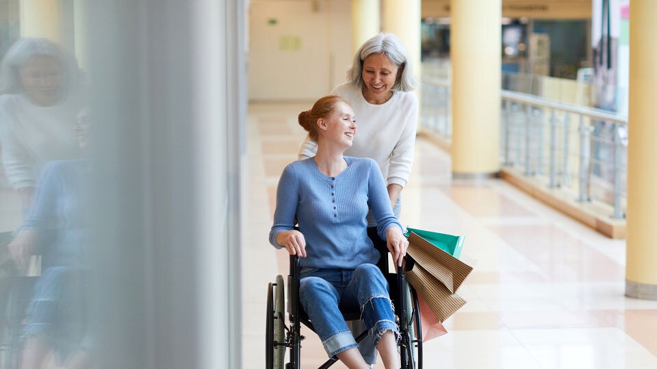 older female pushing younger female in a wheelchair while carrying shopping bags through a retail shopping mall