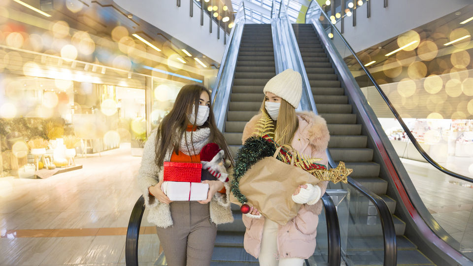 two young women dressed in winter clothes and medical masks descending a retail shopping mall elevator carrying holiday purchases