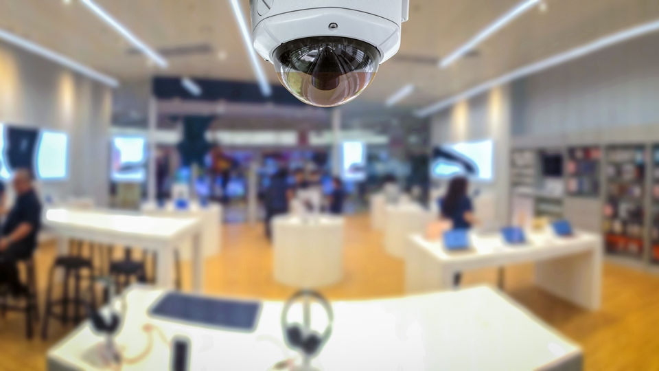 wide angle view of inerior of electronics retail store with shoppers out of focus and ceiling mounted computer vision surveillance camera