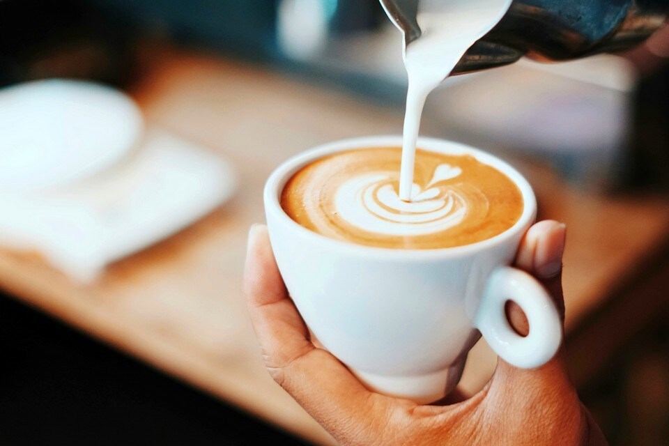 baristas hand pouring frothed milk into an artistic design in a white ceramic coffee cup