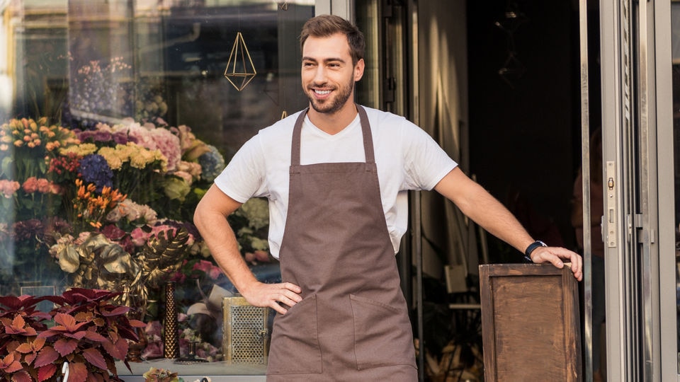 smiling retail shopkeeper wearing brown apron standing outside a store