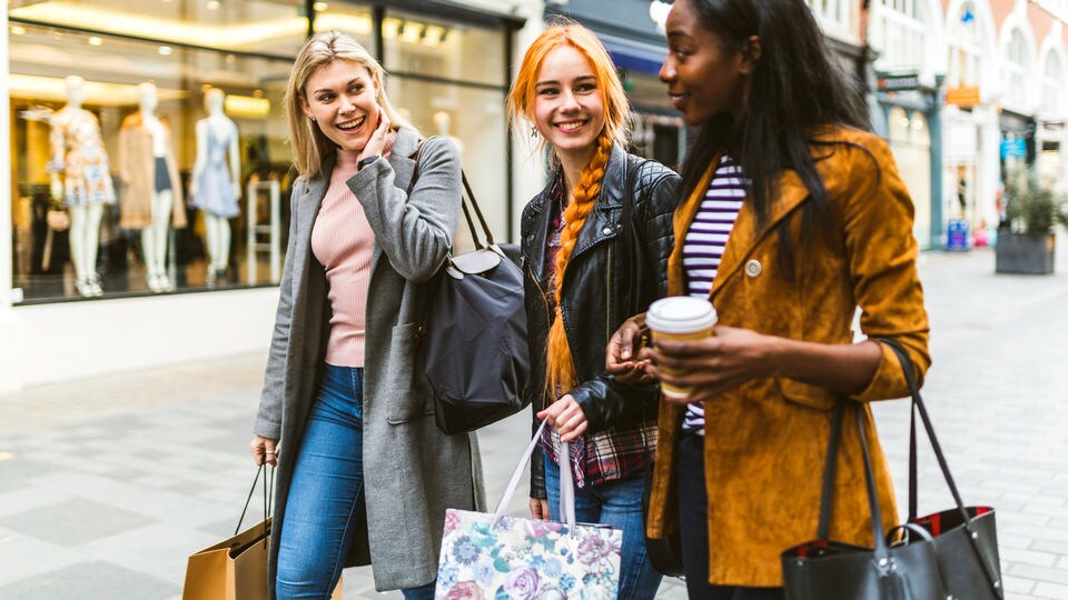 3 female shoppers chatting and carrying shopping bags while walking down the street in a retail shopping district