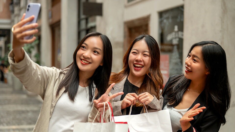 three young female shoppers carrying shopping bags outside retail store taking selfie on smartphone
