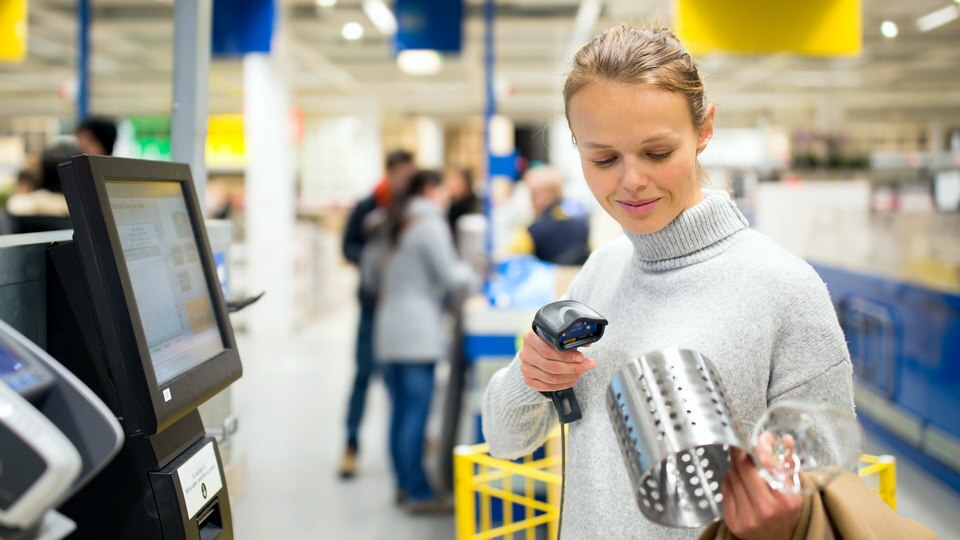 female shopper scans a purchase at the self checkout of a busy retail store with other customers in the background